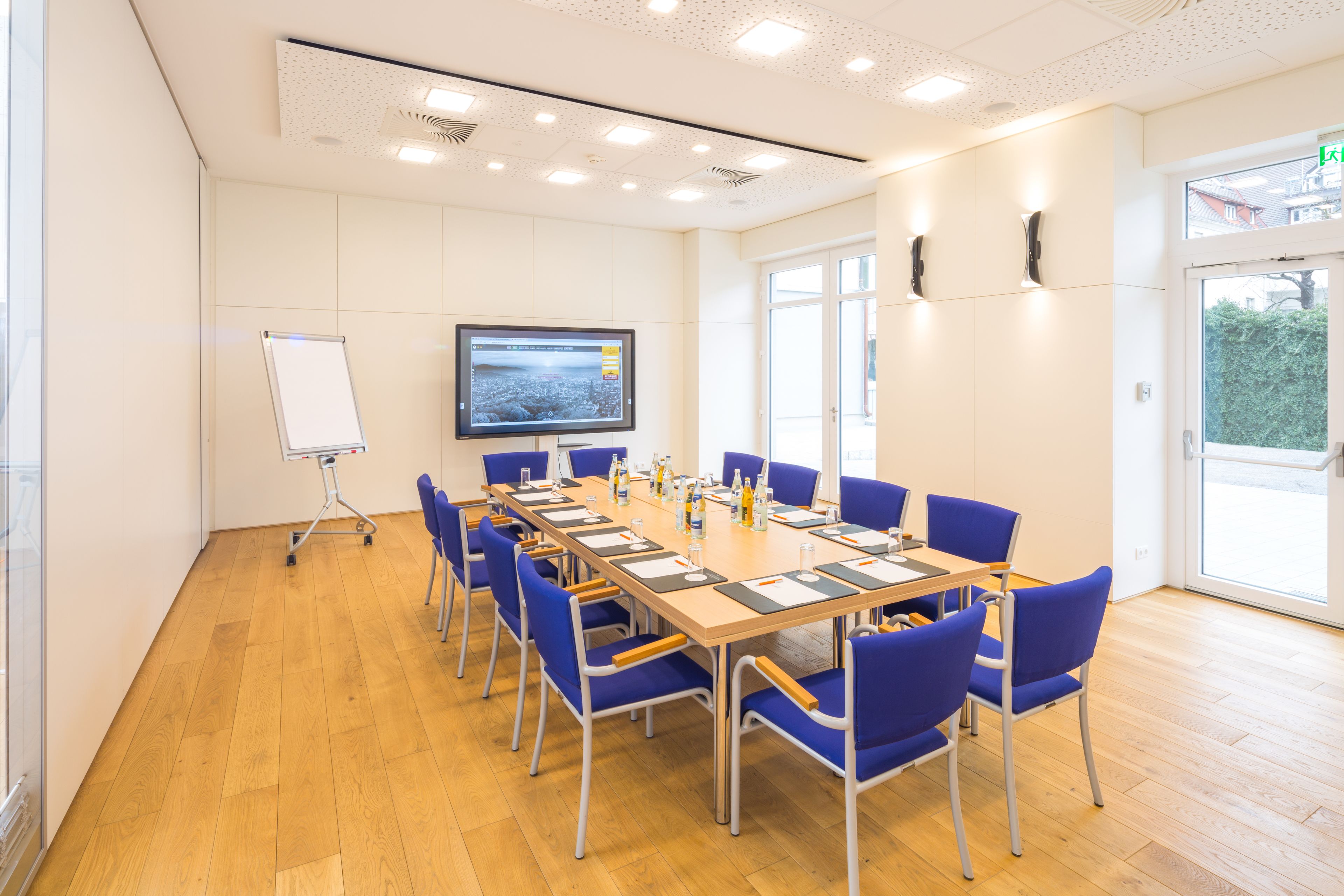 Une salle de conférence avec une longue table, des chaises bleues, un tableau blanc et un grand écran.