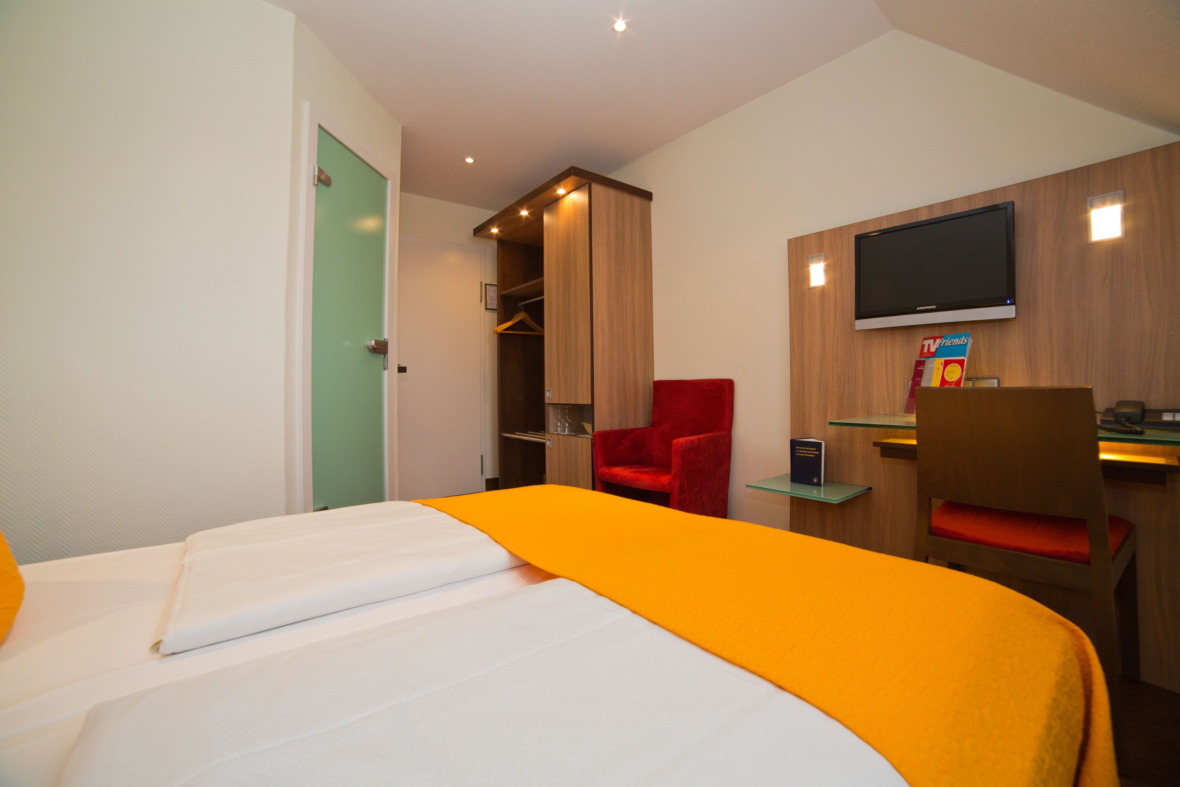A hotel room with a bed, a red chair, a desk, a television, and a wardrobe.