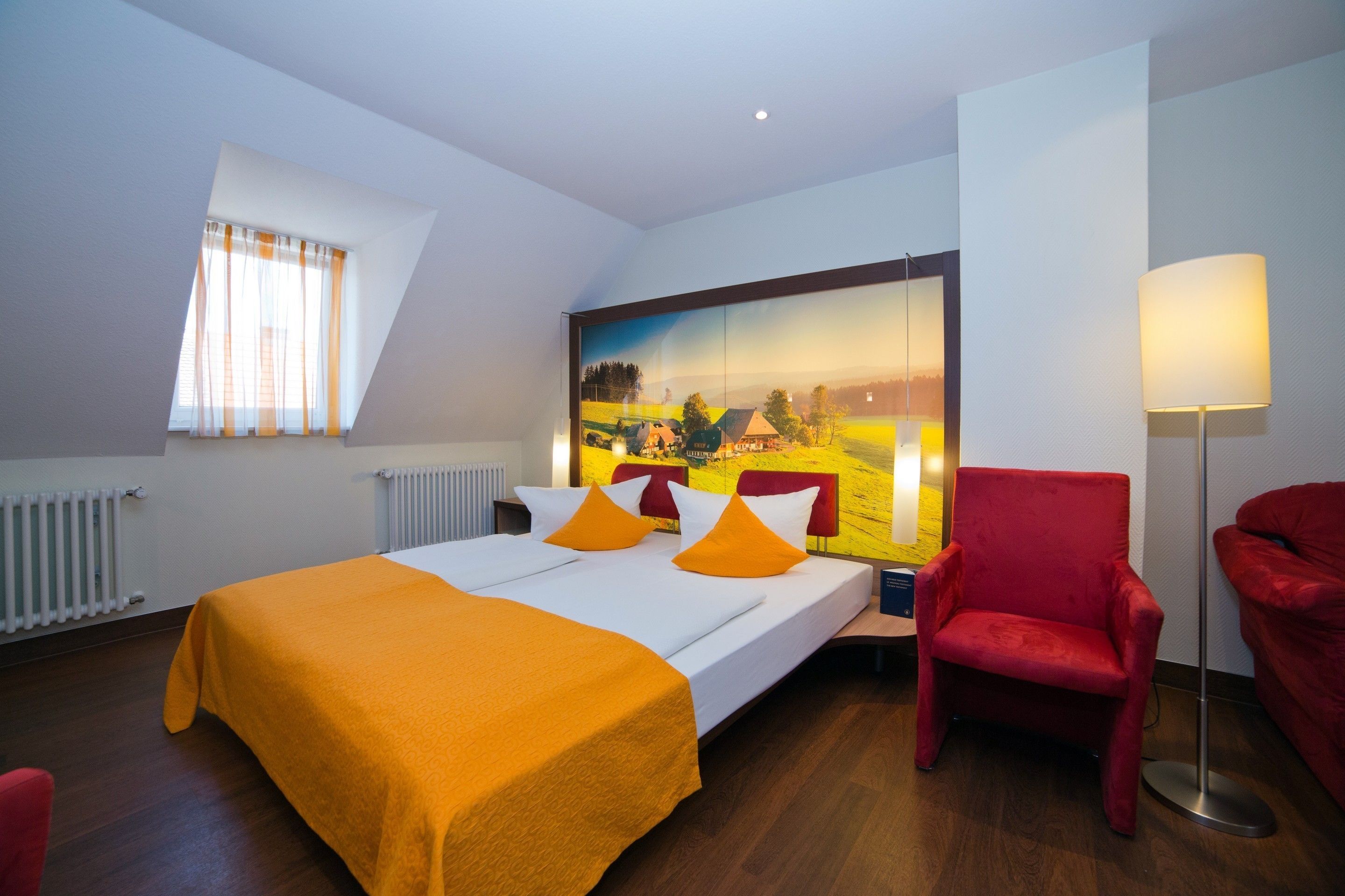 A hotel room with a bed, two red chairs, a large painting on the wall, and a window with curtains.