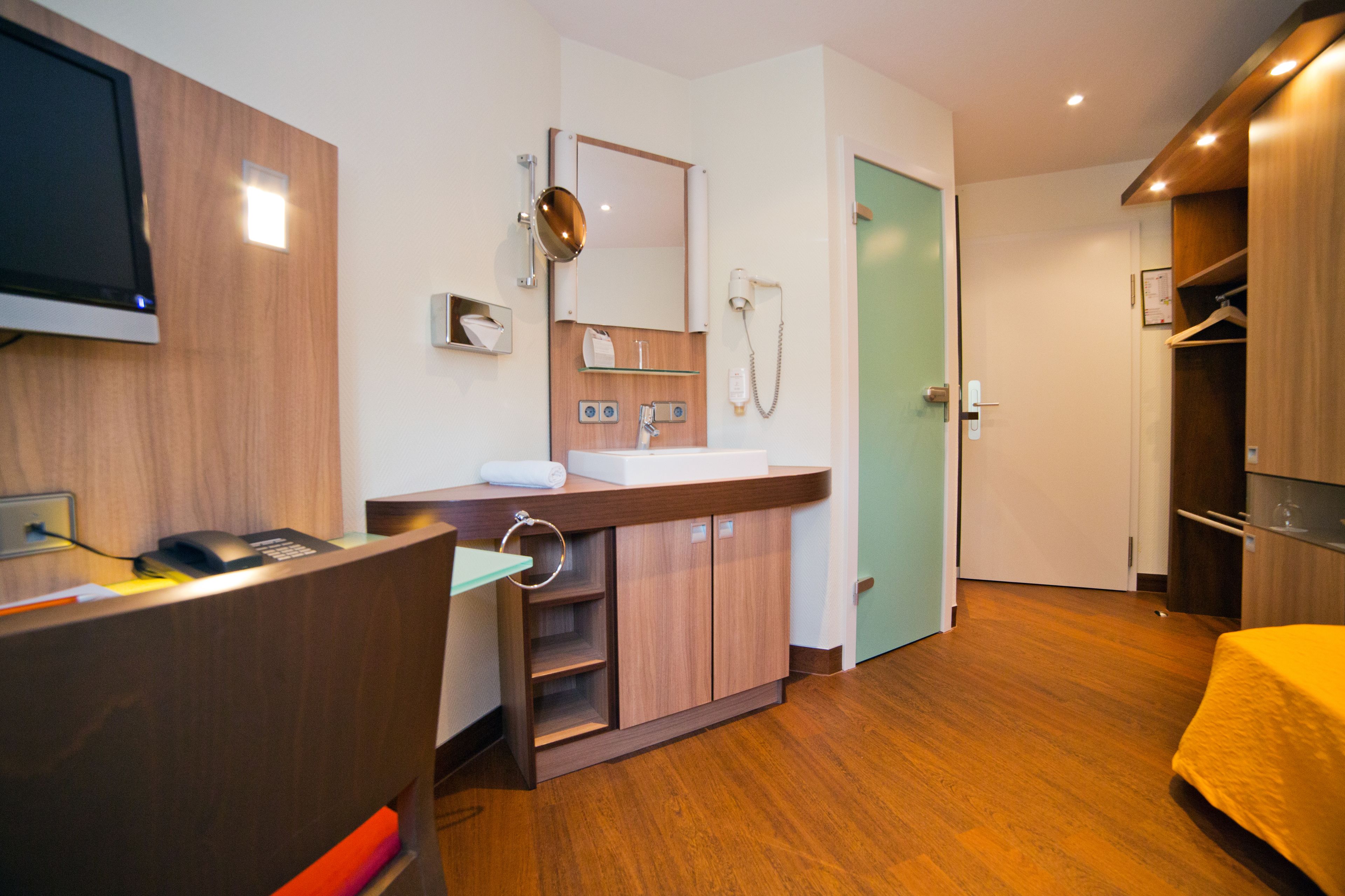 A modern hotel room with a wooden floor, a wooden vanity with a mirror, a sink, and a shower. There is a bed with a yellow bedspread and a wooden wardrobe.