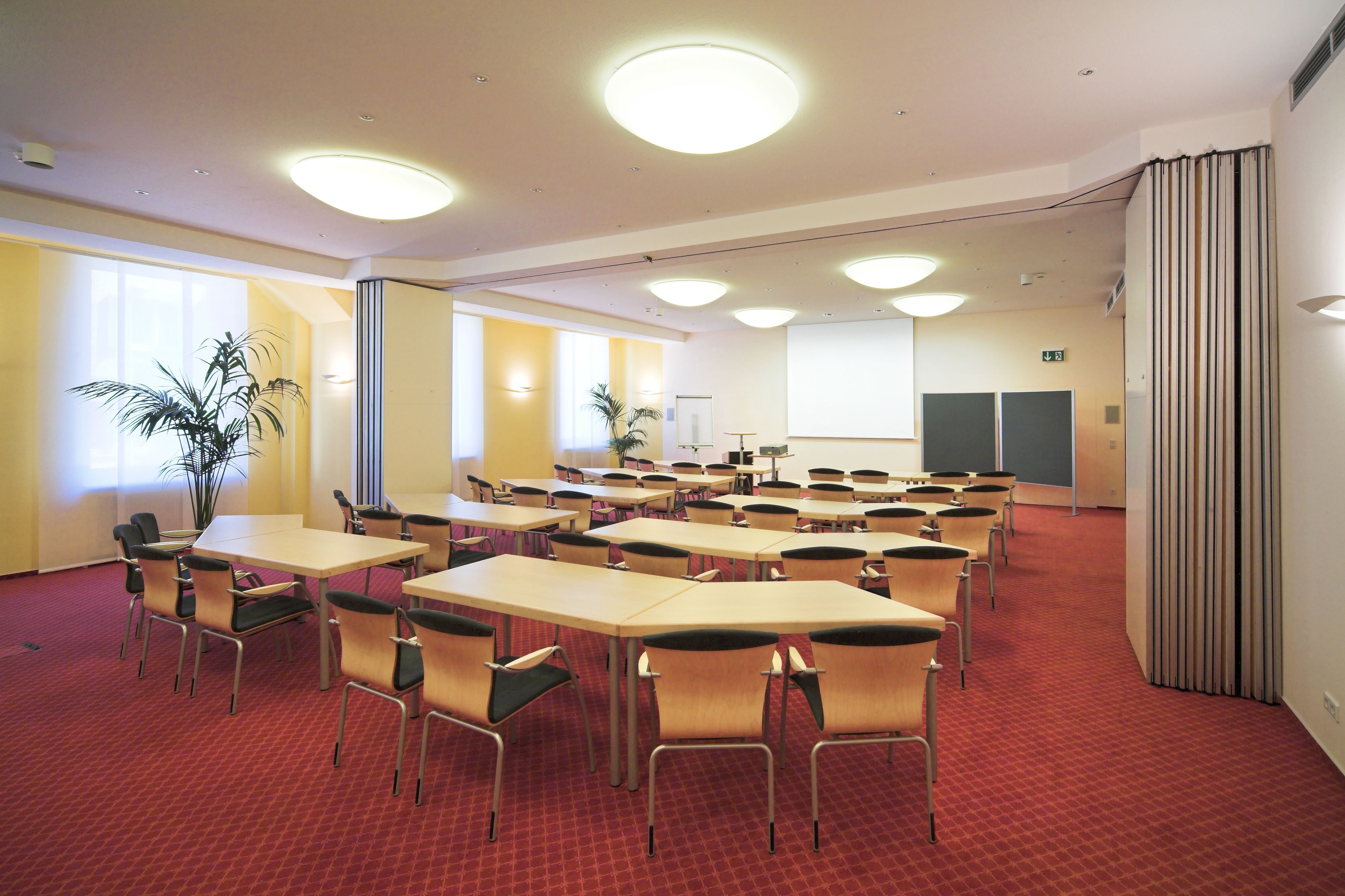 Une salle de conférence équipée de plusieurs tables et chaises, d’un écran de projection et d’une grande fenêtre avec des stores.