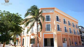 The exterior of the Los Itzaes Hotel with a palm tree in the foreground, a clear blue sky, and a street in front.