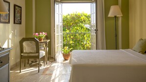 Habitación de hotel con cama, una silla, una mesa con planta en maceta y vista a la vegetación a través de una ventana abierta.