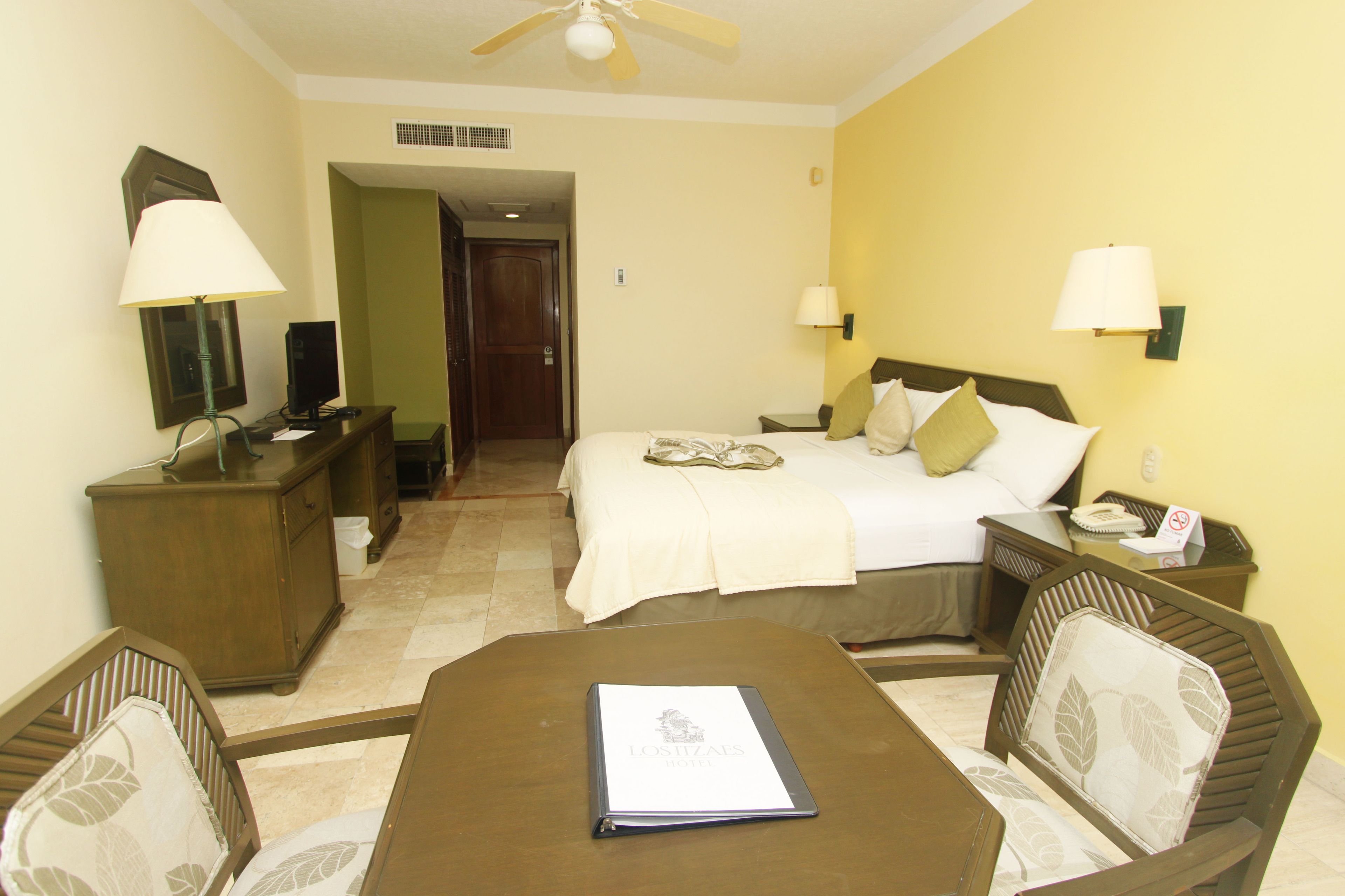 Une chambre d’hôtel comprenant un lit, des tables de chevet, un bureau avec une chaise, un ventilateur de plafond et donnant sur le couloir.