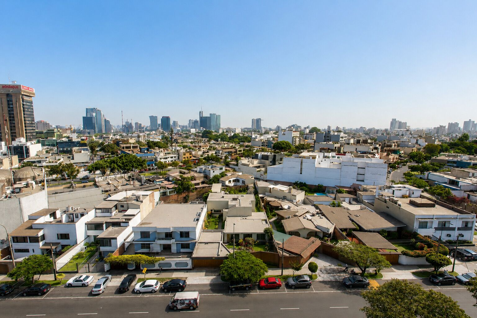 A residential area with a mix of modern and older buildings, a clear sky, and a parking lot with several cars.