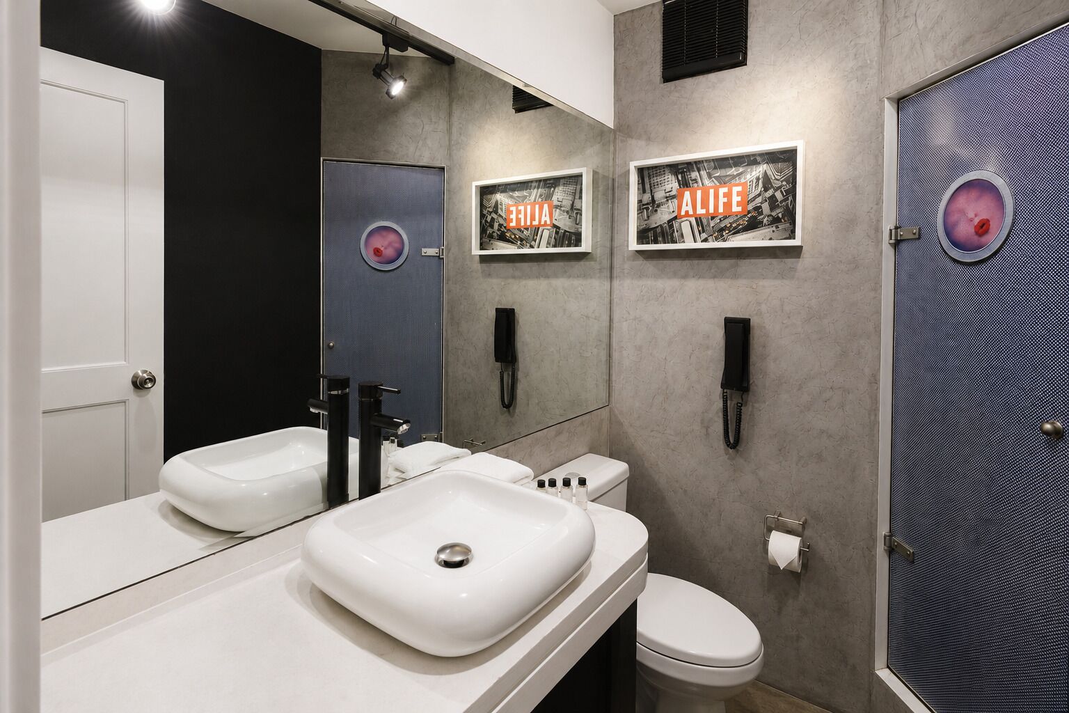 Un baño moderno con un lavamanos blanco, dos grifos negros, un inodoro y un espejo. Hay dos cuadros en la pared, uno con la palabra 'ALIFE' y otro con un paisaje urbano.