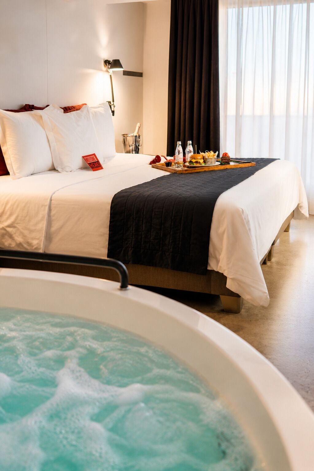 Habitación de hotel con cama, un servicio de comidas, bañera de hidromasaje y mesita de noche con lámpara.