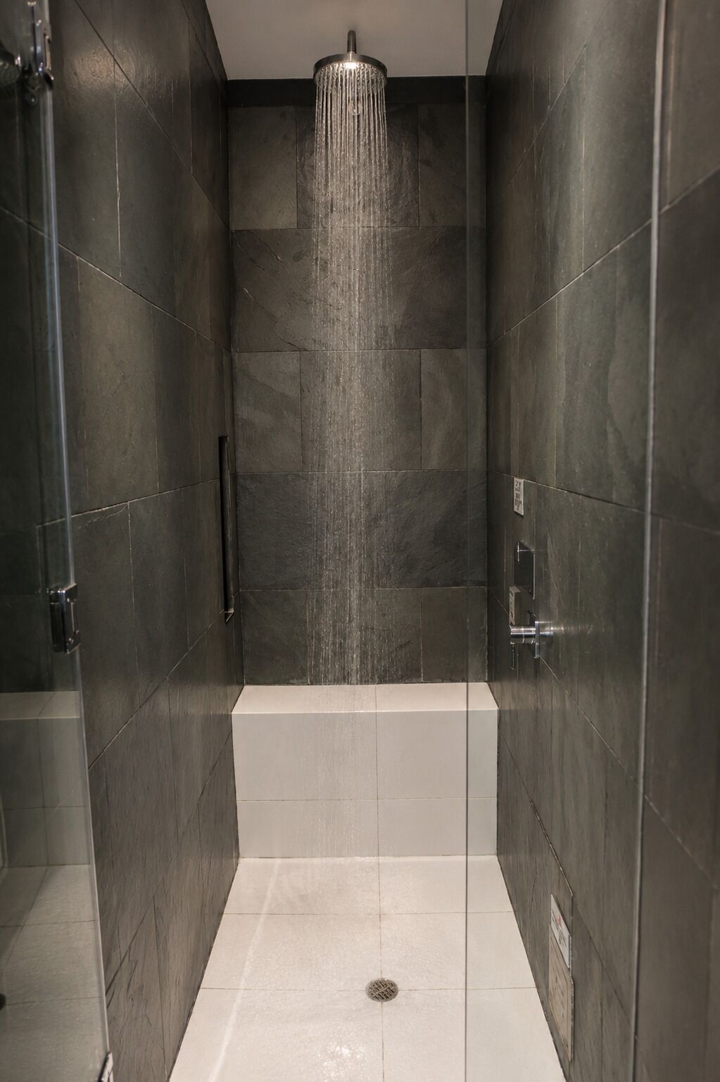 A modern shower with a rain showerhead, a bathtub, and dark tiled walls.