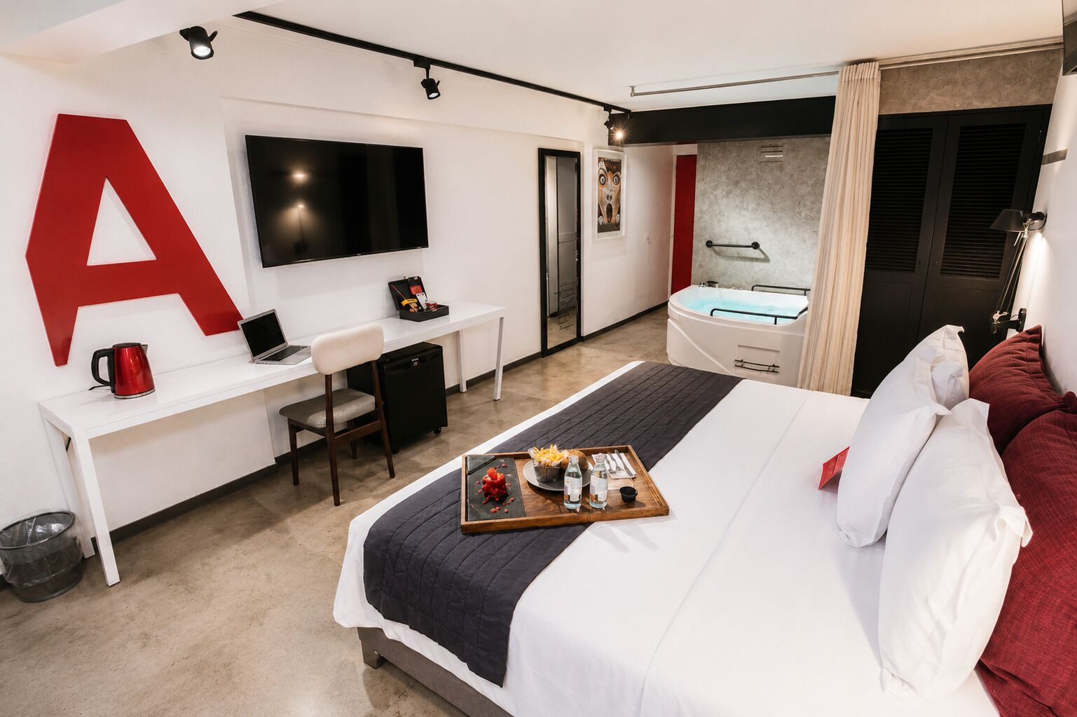 A modern hotel room with a large bed, a desk with a laptop, a TV, a hot tub, and a large red letter 'A' on the wall.