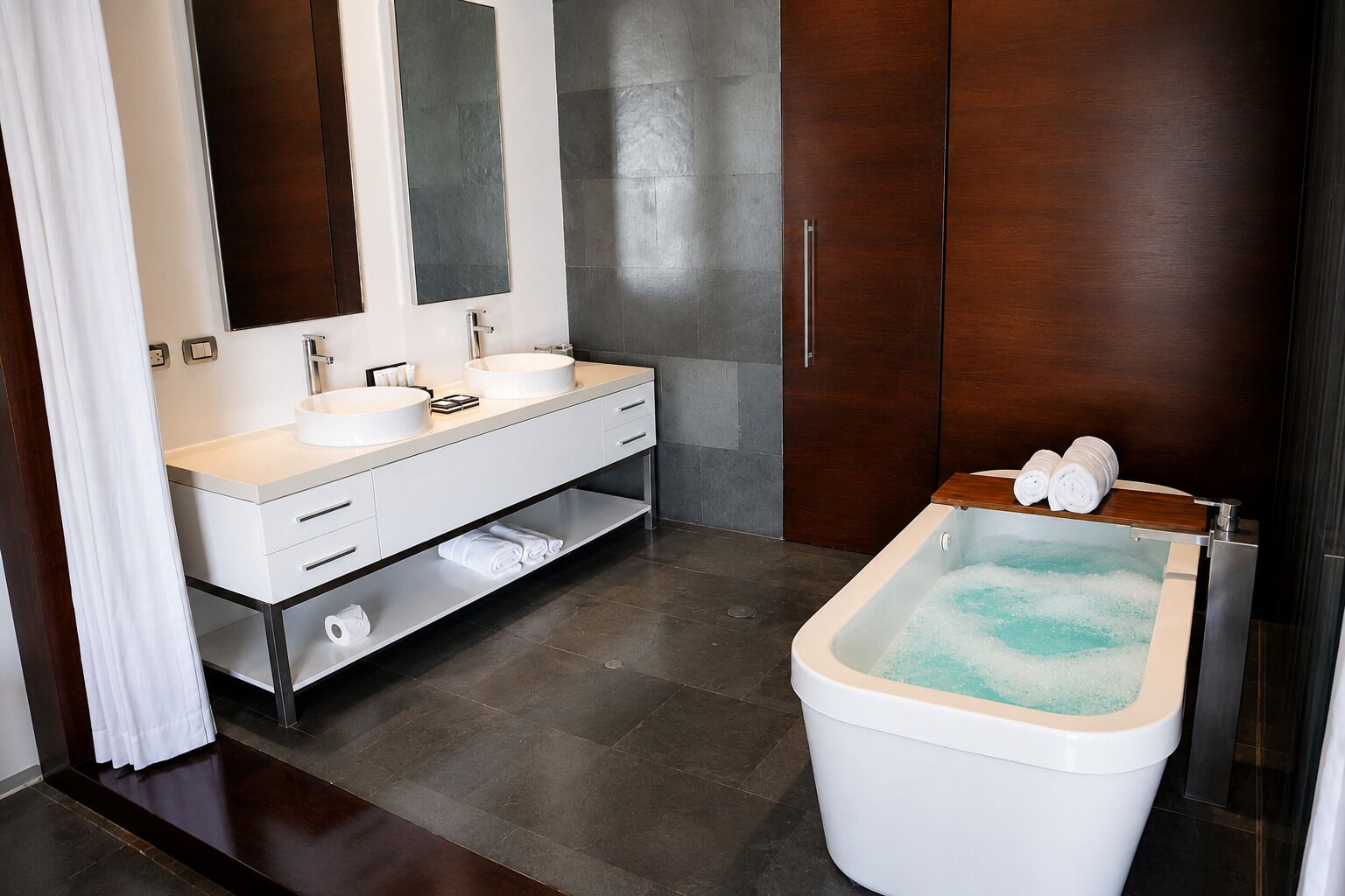A modern bathroom with a double sink vanity, a large mirror, a wooden cabinet, and a standalone bathtub with towels.