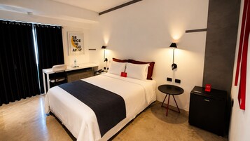 Una habitación de hotel con cama, mesita de noche, lámpara y un pequeño armario negro.