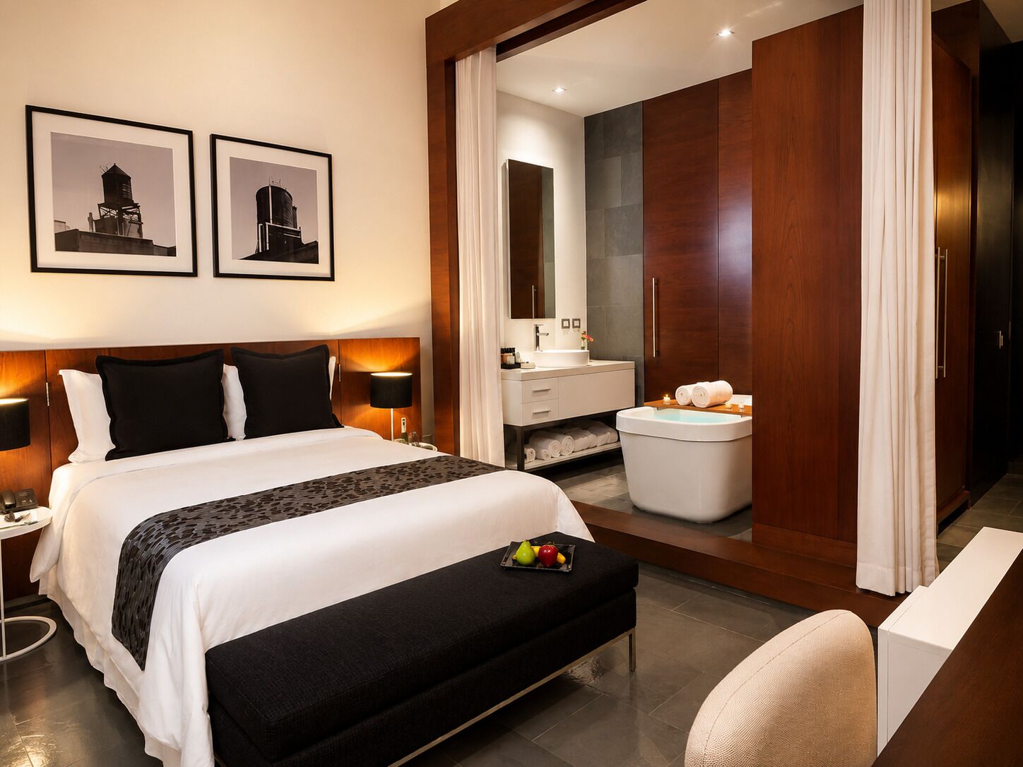 Una habitación de hotel con cama, lámparas de noche, mesita de noche, un banco con fruta, y un baño con lavamanos, espejo e inodoro.