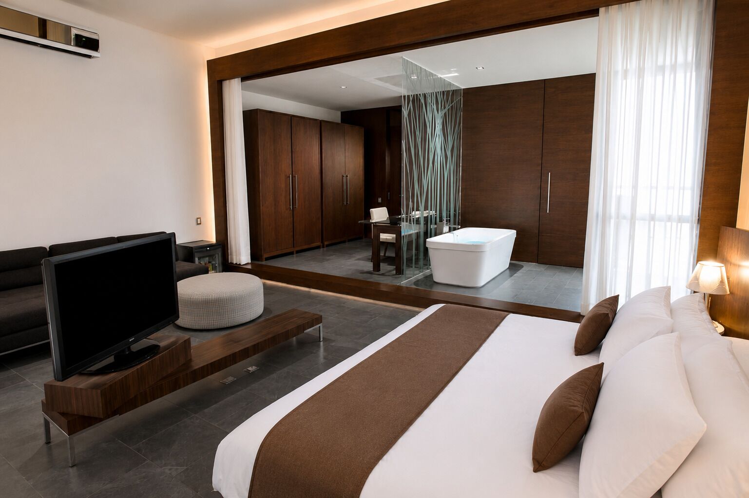 A modern hotel room with a large bed, a flat-screen TV, a seating area, and a bathroom visible through a glass partition.