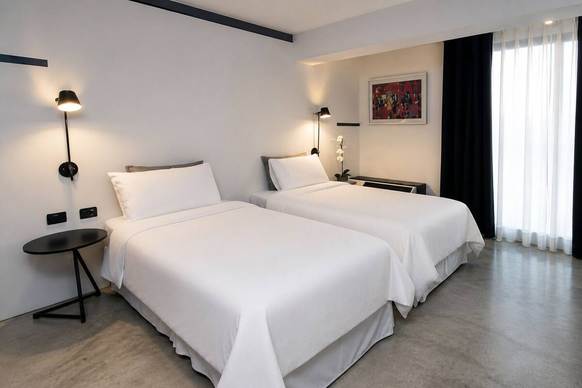 Habitación de hotel con dos camas, una mesita, una silla y un cuadro en la pared.