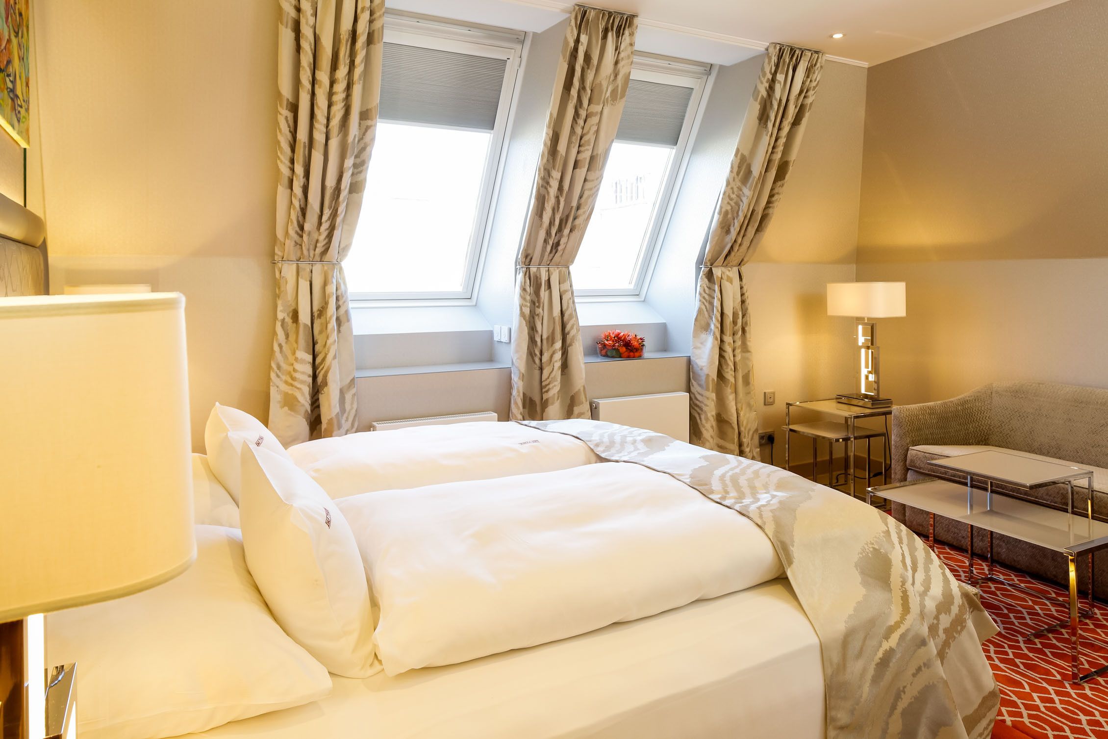 A hotel room with a large bed, a desk, a chair, and a sofa. There are two windows with curtains, a lamp, and a small table with a vase of flowers.