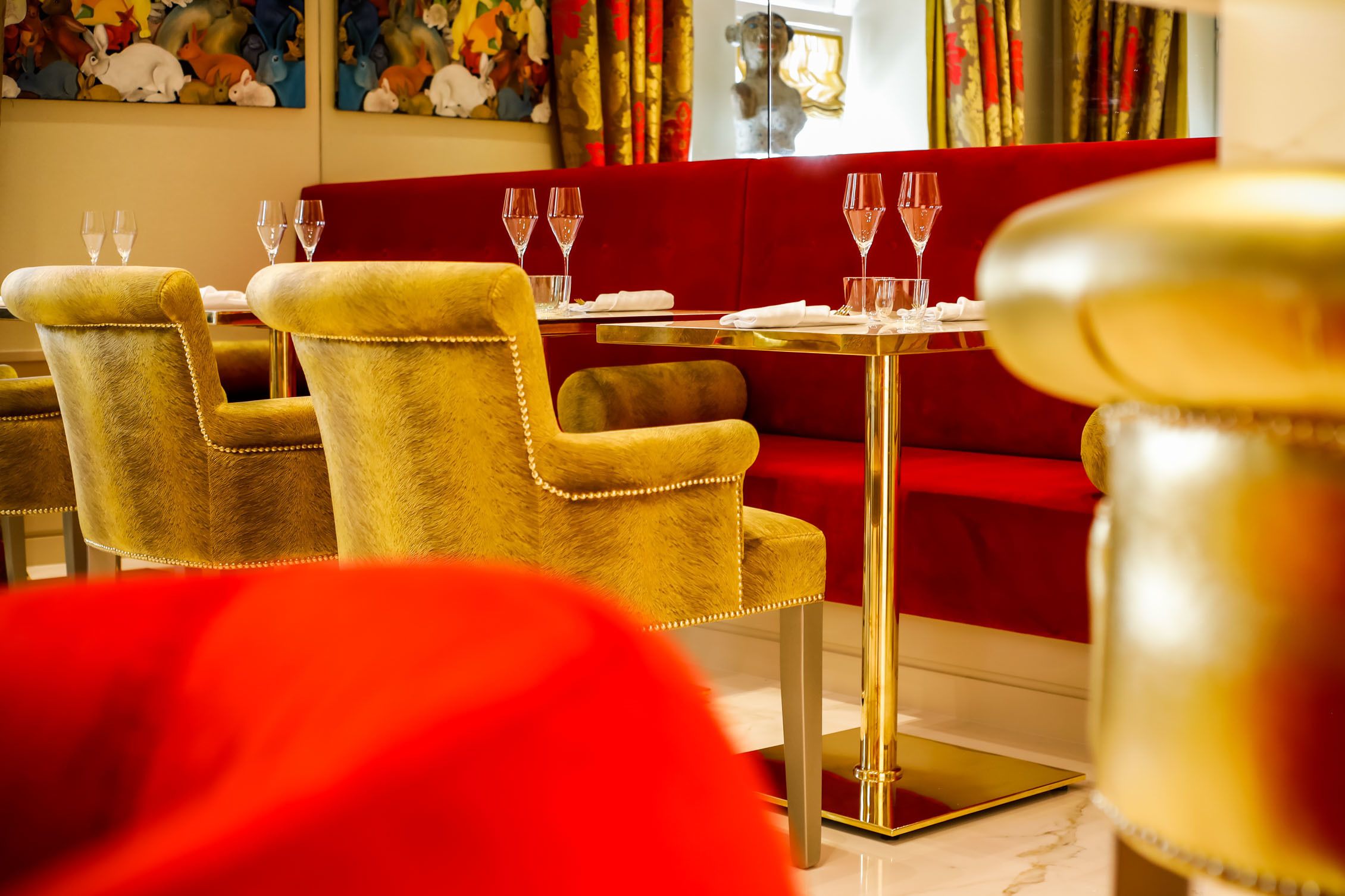 Un comedor con sillas de color dorado y tapicería mullida, un sofá rojo y una mesa dorada dispuesta con copas de vino.