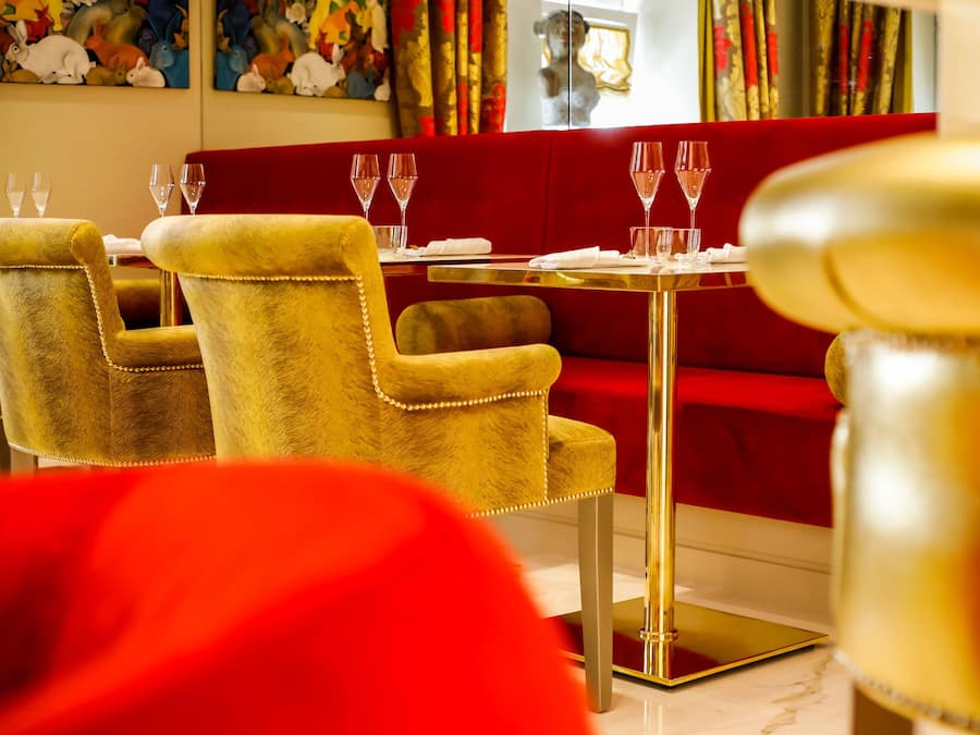 Un comedor con sillas de color dorado y tapicería mullida, un sofá rojo y una mesa dorada dispuesta con copas de vino.