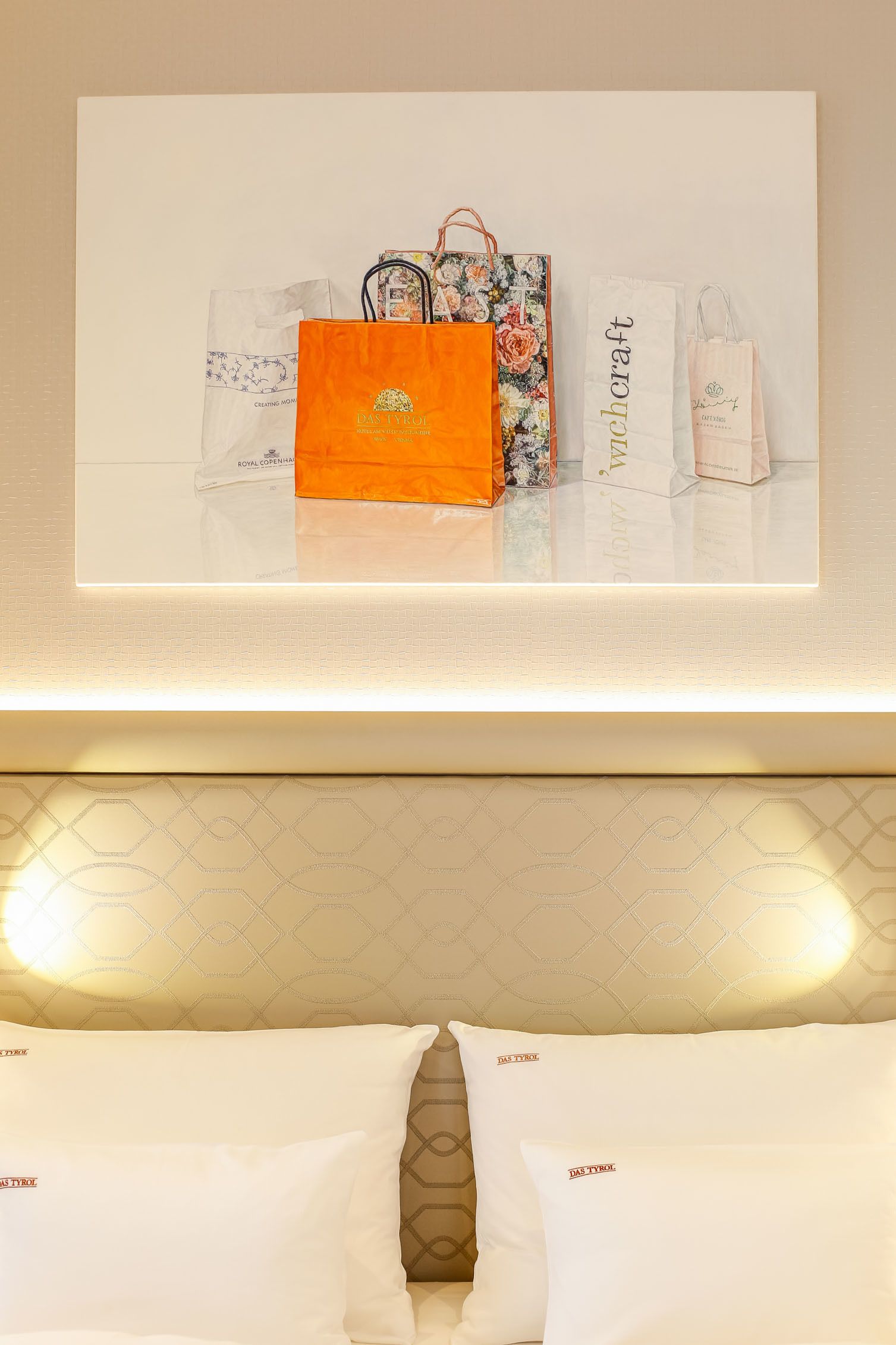 A framed artwork of shopping bags on a wall, a bed with white pillows, and a patterned headboard.