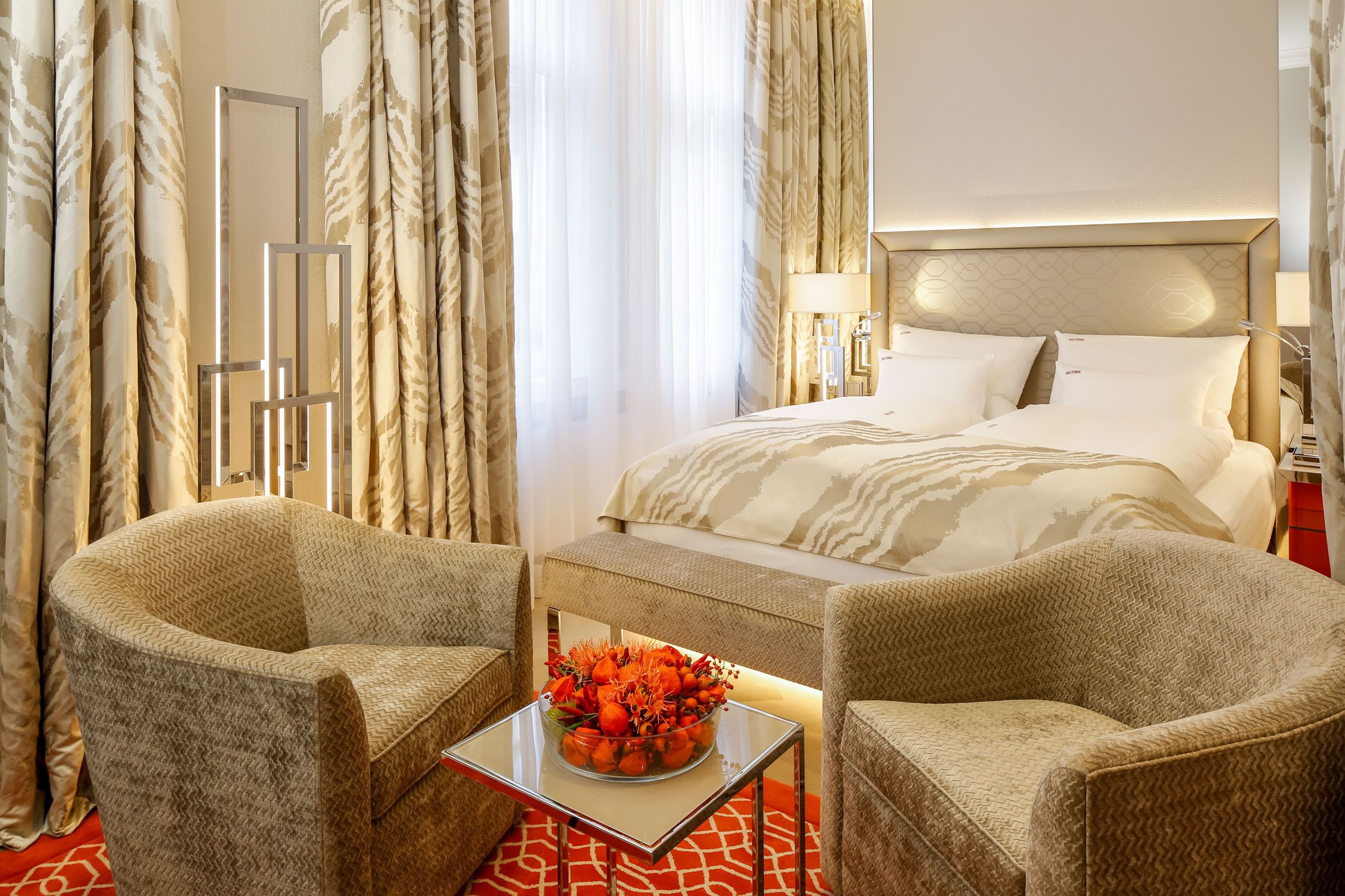 Ein Hotelzimmer mit einem Bett, zwei Sesseln, einem kleinen Tisch mit Blumen und einem Fenster mit Vorhängen.