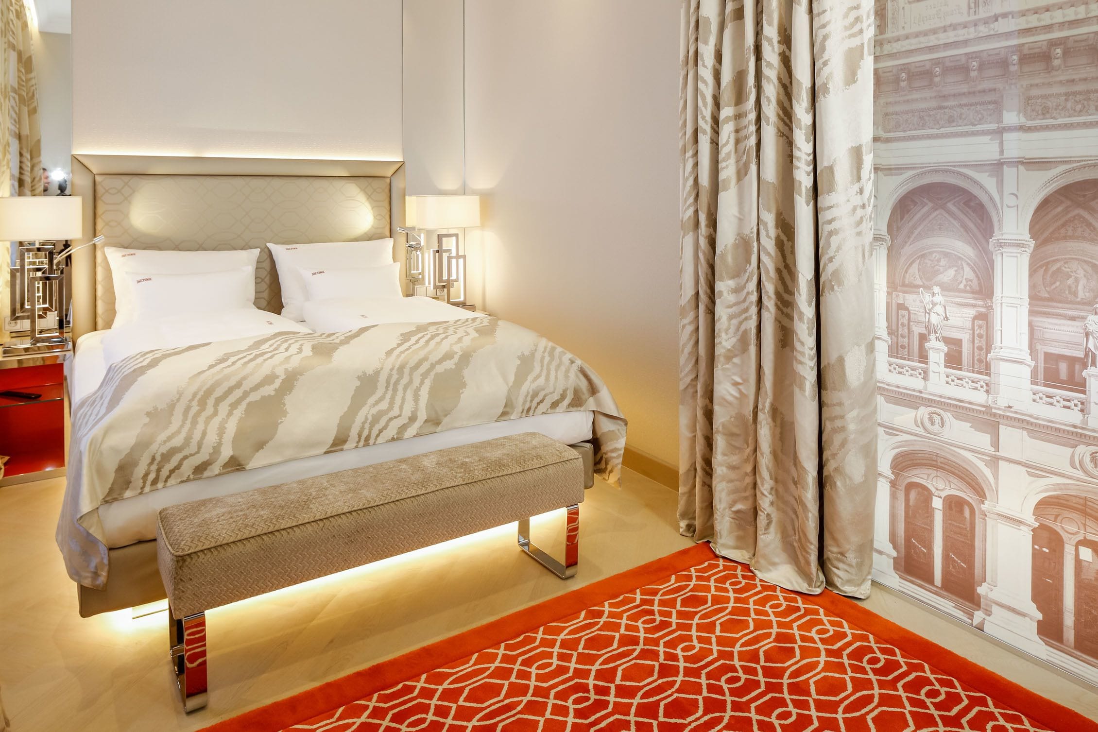A hotel room with a bed, bedside lamps, a bench, a patterned rug, and a view of a building through curtains.