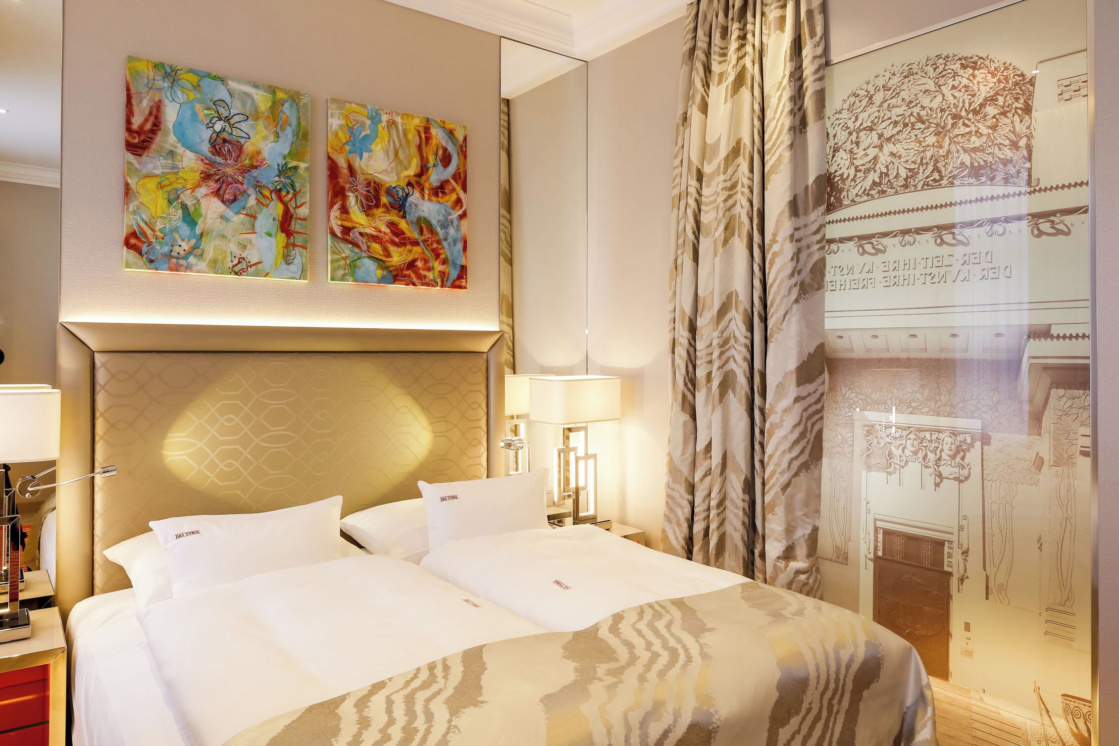 A hotel room with a large bed, two bedside tables, a headboard with a geometric pattern, and two abstract paintings on the wall.