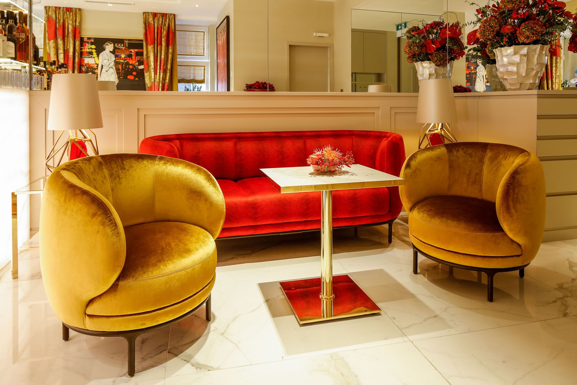 Een moderne woonkamer met een rode velourse bank, twee gouden fauteuils en een kleine vierkante tafel met een rode voet.