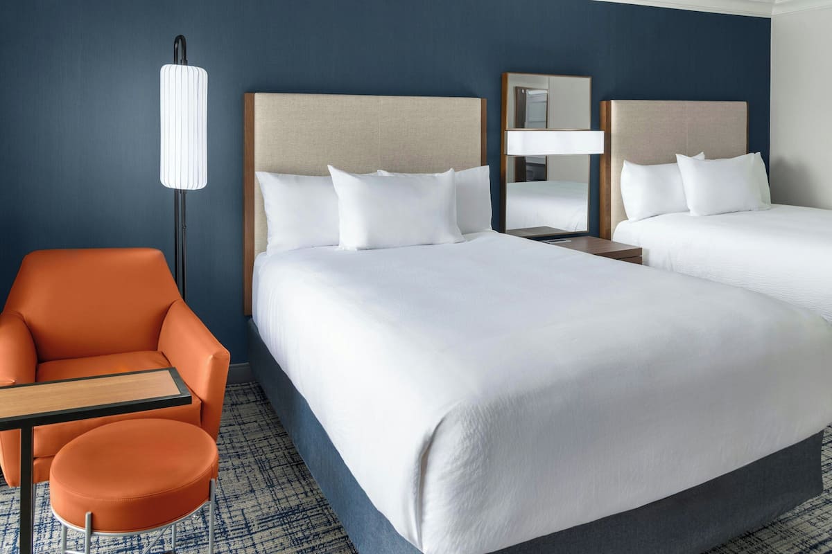 A hotel room with two beds, a nightstand, a lamp, an orange chair, and a small table.
