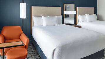 Habitación de hotel con dos camas, una mesita de noche, una lámpara, un sillón naranja y una pequeña mesa.