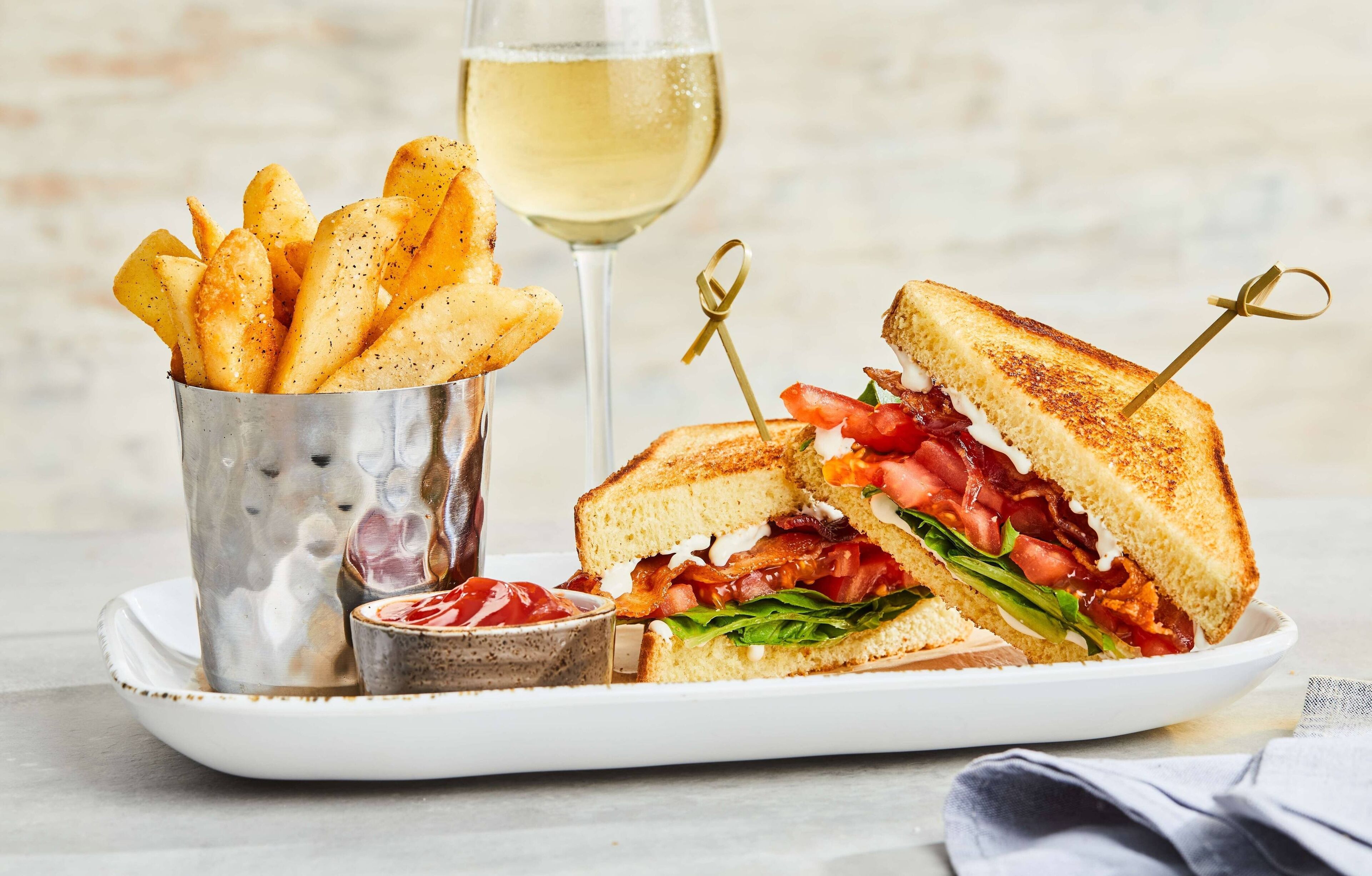A plate with a grilled sandwich, a glass of white wine, and a container of fries.