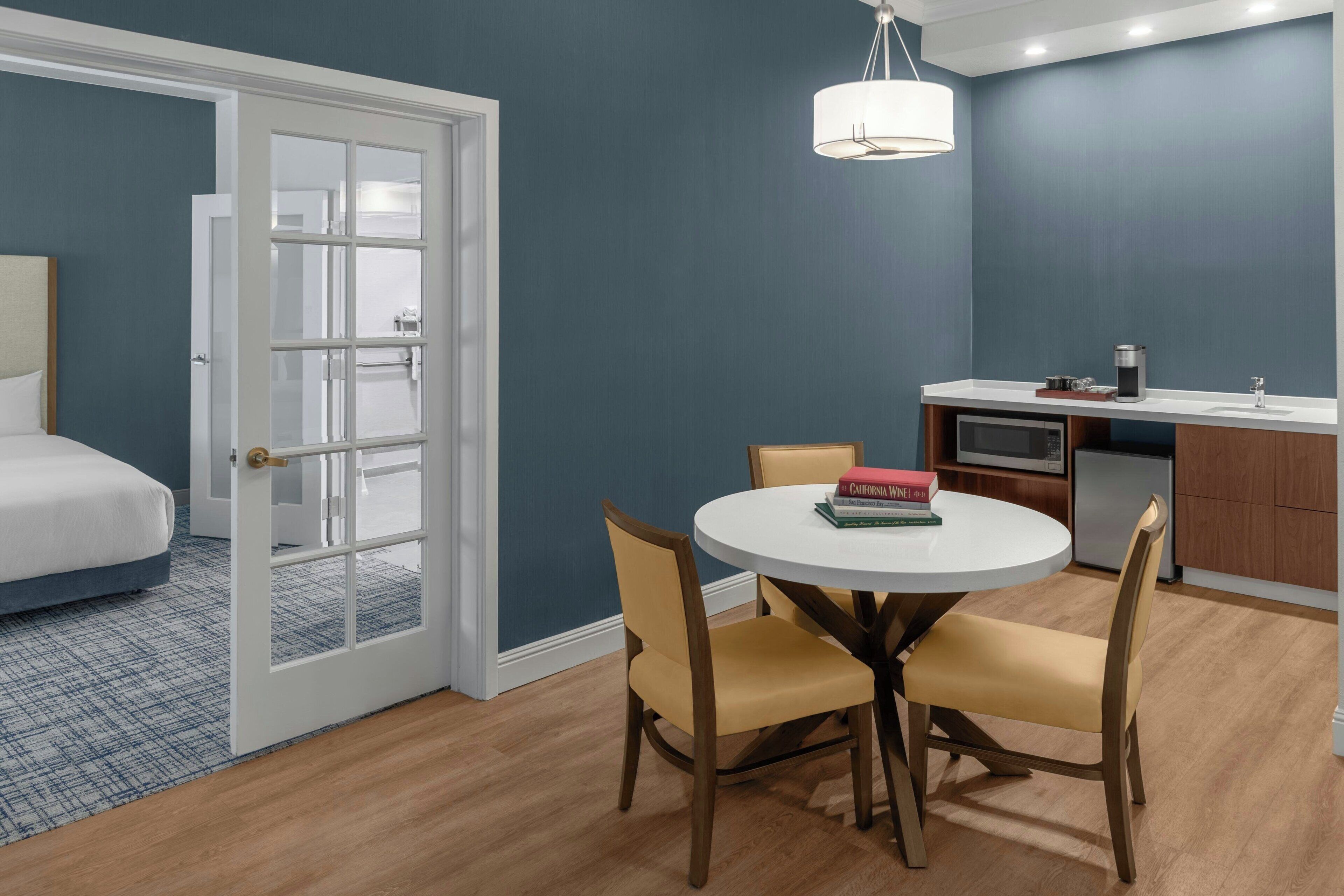 Habitación de hotel compacta con cama, zona de comedor con una mesa redonda y sillas, kitchenette y puerta que da a otra habitación.