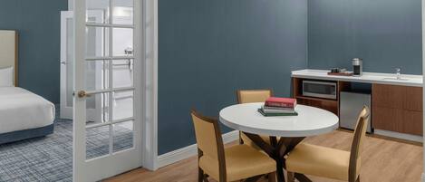 Habitación de hotel compacta con cama, zona de comedor con una mesa redonda y sillas, kitchenette y puerta que da a otra habitación.