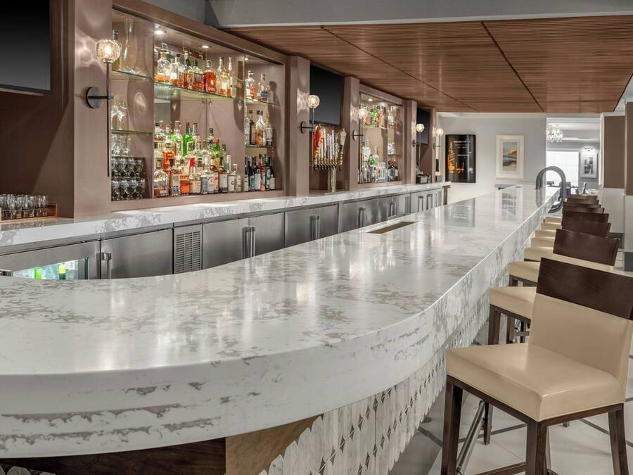 Un área de bar moderna con una barra curva de mármol, varias estanterías llenas de botellas de licores y taburetes de bar.