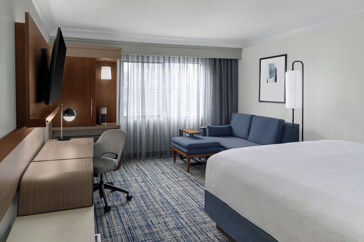 A hotel room with a bed, a desk, a chair, a TV, a lamp, and a window with curtains.