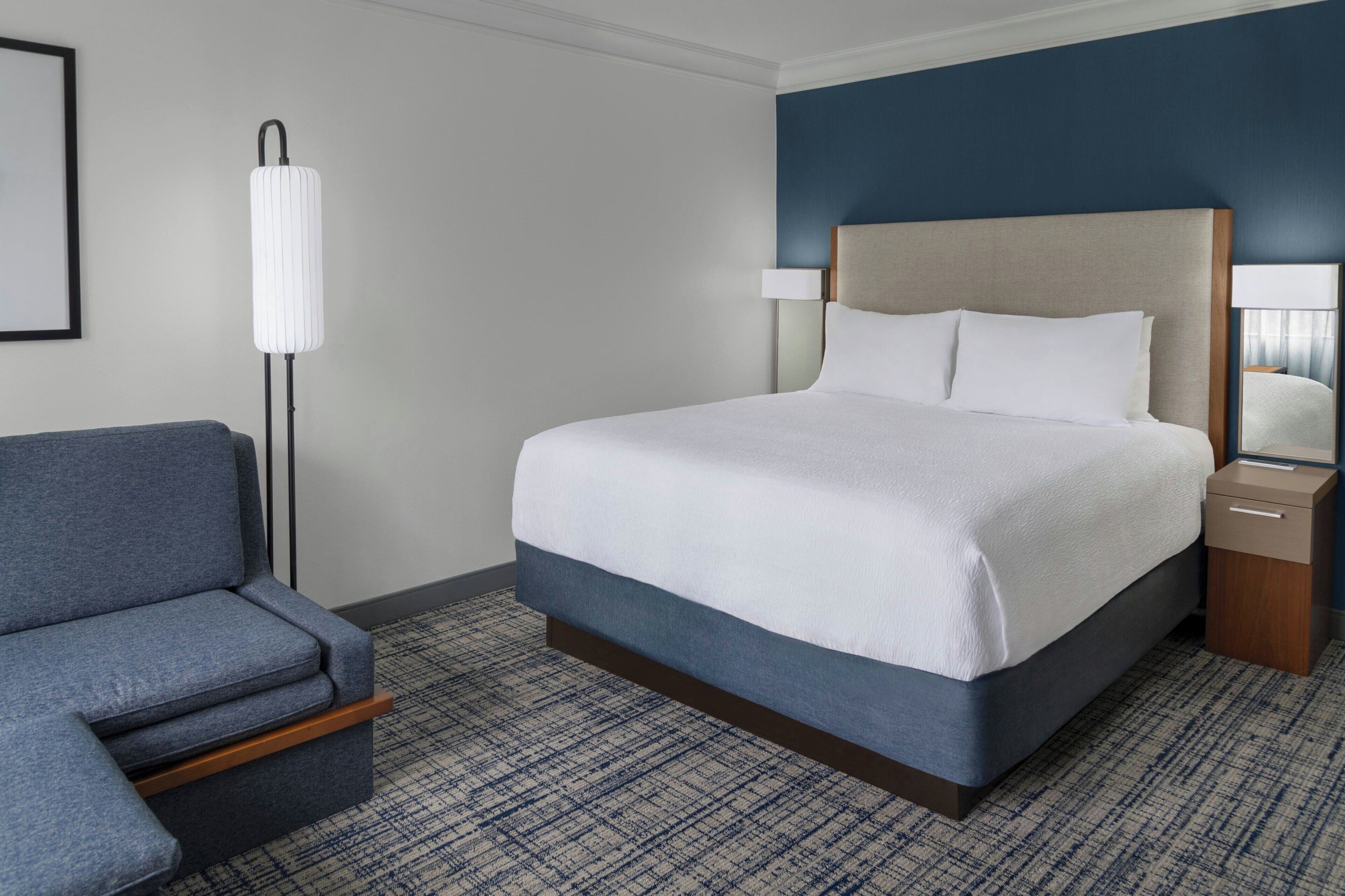 Une chambre d’hôtel avec un lit, une chaise, une table de chevet et une lampe sur pied.