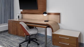 Habitación de hotel moderna con un escritorio de madera, una silla de oficina gris, un televisor de pantalla plana montado en la pared y una mesita de noche con cajones.