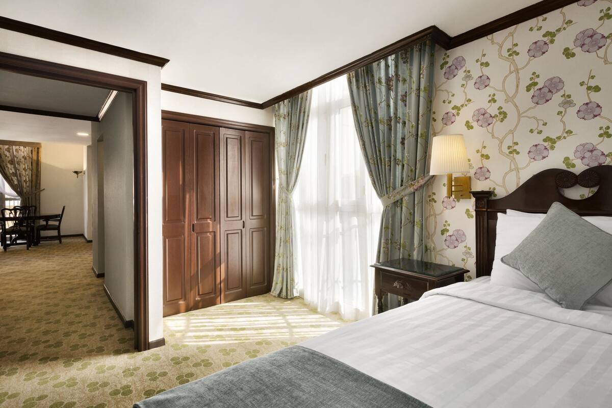 Une chambre d’hôtel comprenant un lit, une table de chevet, une lampe, une armoire en bois et un bureau avec des chaises.
