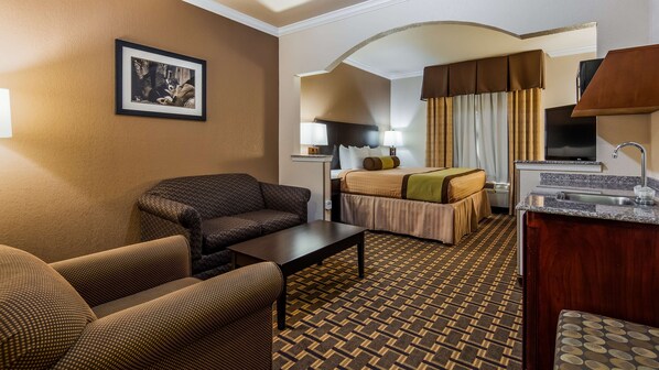 Ein Hotelzimmer mit einer Couch, einem Bett, einem Schreibtisch, einem Spiegel, einem Bild an der Wand und einer Küchenzeile mit Spüle und Fernseher.
