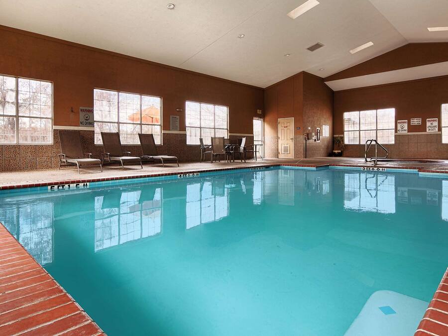 An indoor swimming pool with clear water, surrounded by lounge chairs and tables, large windows, and warning signs.