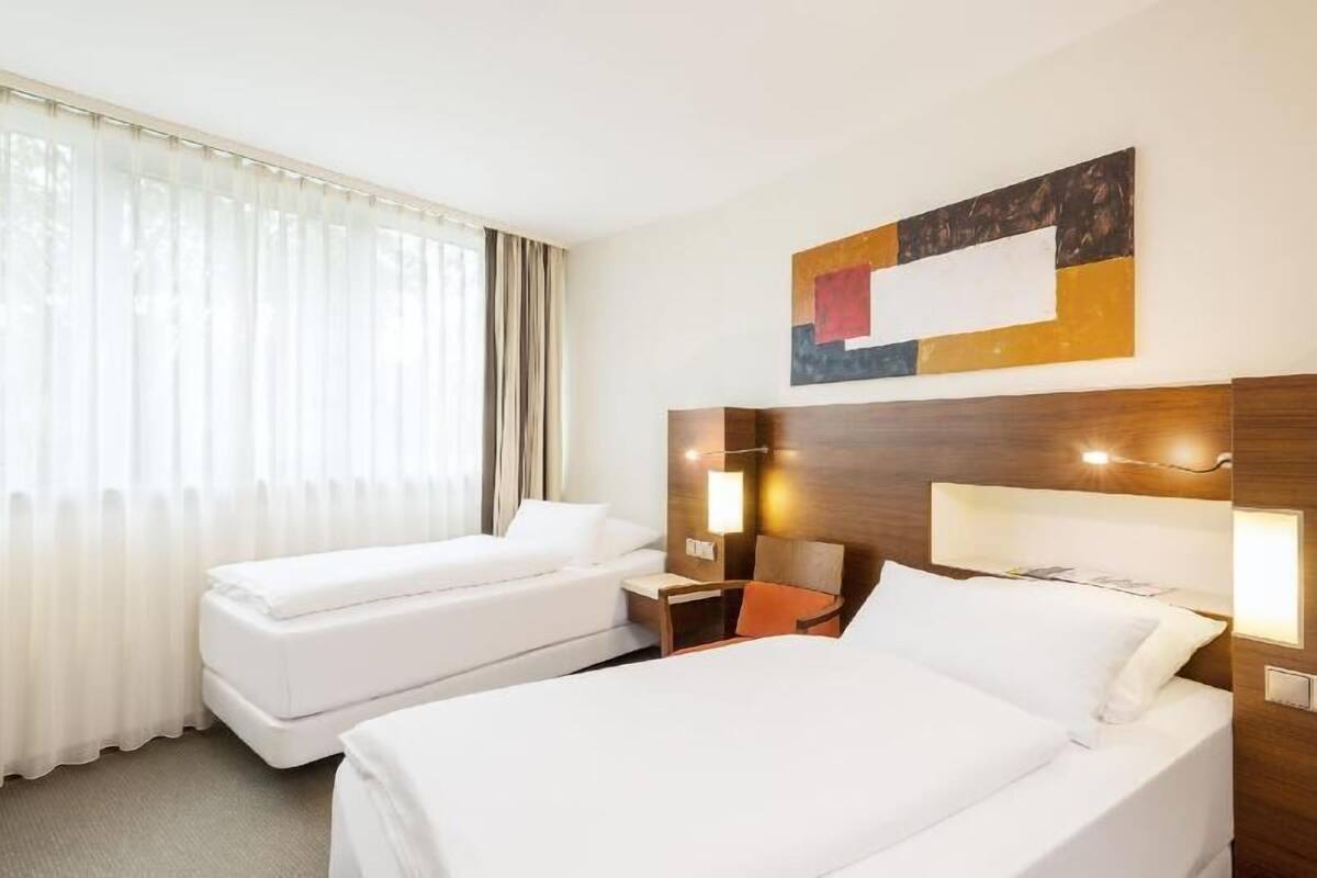 Habitación de hotel con dos camas, cabecera de madera, una obra de arte en la pared y una ventana con cortinas translúcidas.