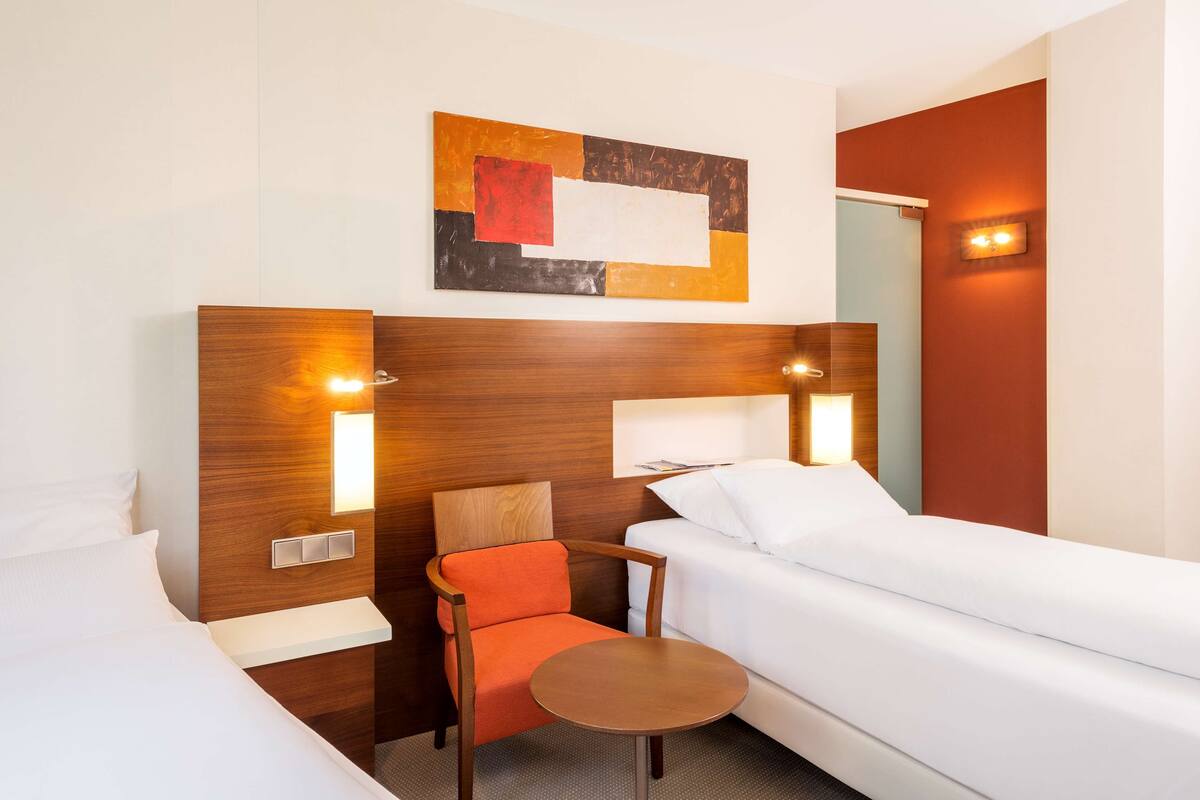 A hotel room with a wooden headboard, a single bed, a small table, a chair, and a wall-mounted artwork.