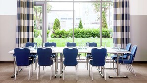 A conference room with a large table and blue chairs, large windows with curtains, and a view of greenery and buildings outside. - NH Frankfurt Niederrad (Frankfurt)