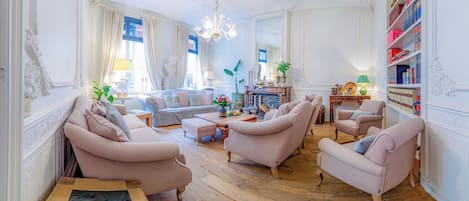 Un salon spacieux comprenant un canapé, des fauteuils, une table basse et une cheminée. On y trouve également des étagères et un lustre.