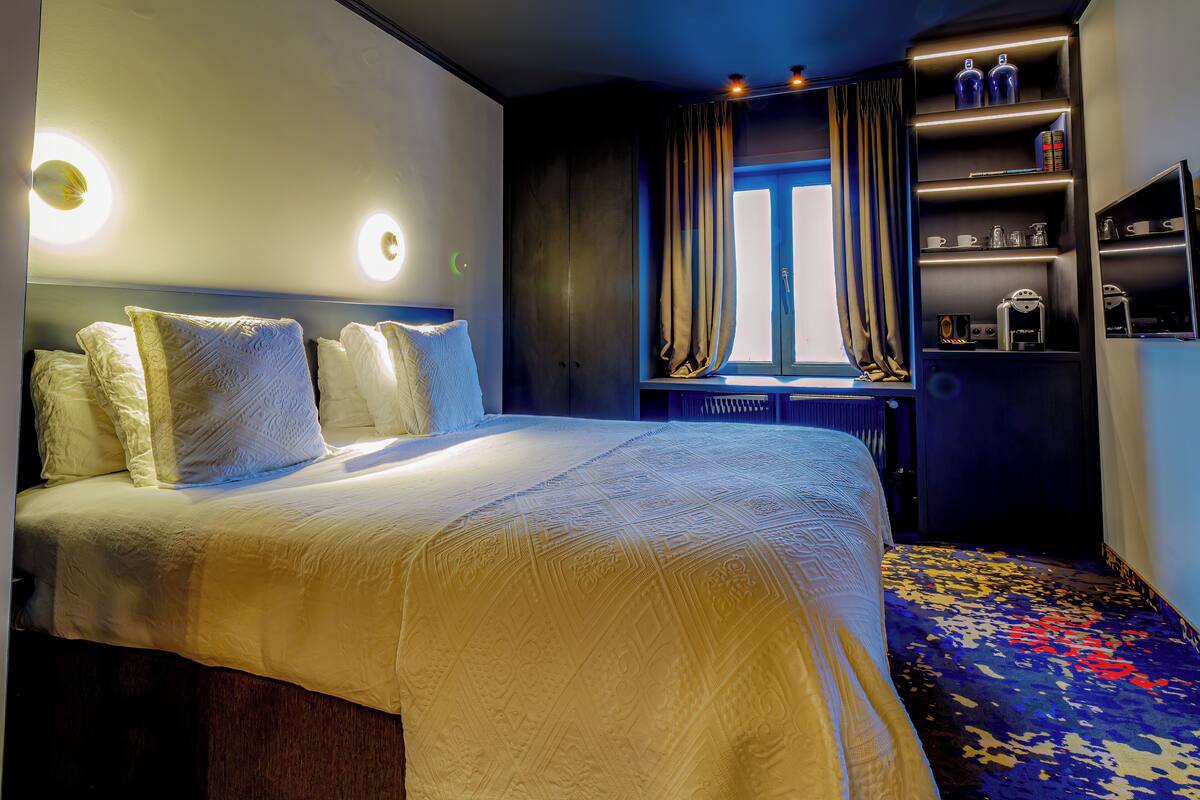 A hotel room with a large bed, a window with curtains, a shelf with toiletries, and a patterned carpet.