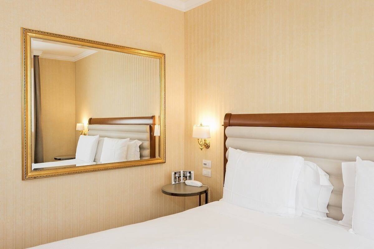 A hotel room with a large mirror, a bed with white linens, a bedside table with a lamp, and a wall-mounted light fixture.