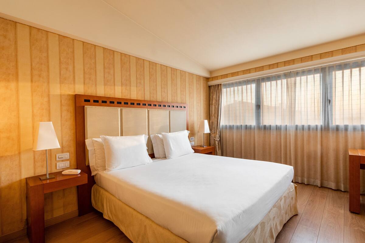 A hotel room with a large bed, wooden panel walls, a nightstand with a lamp, and a window with curtains.