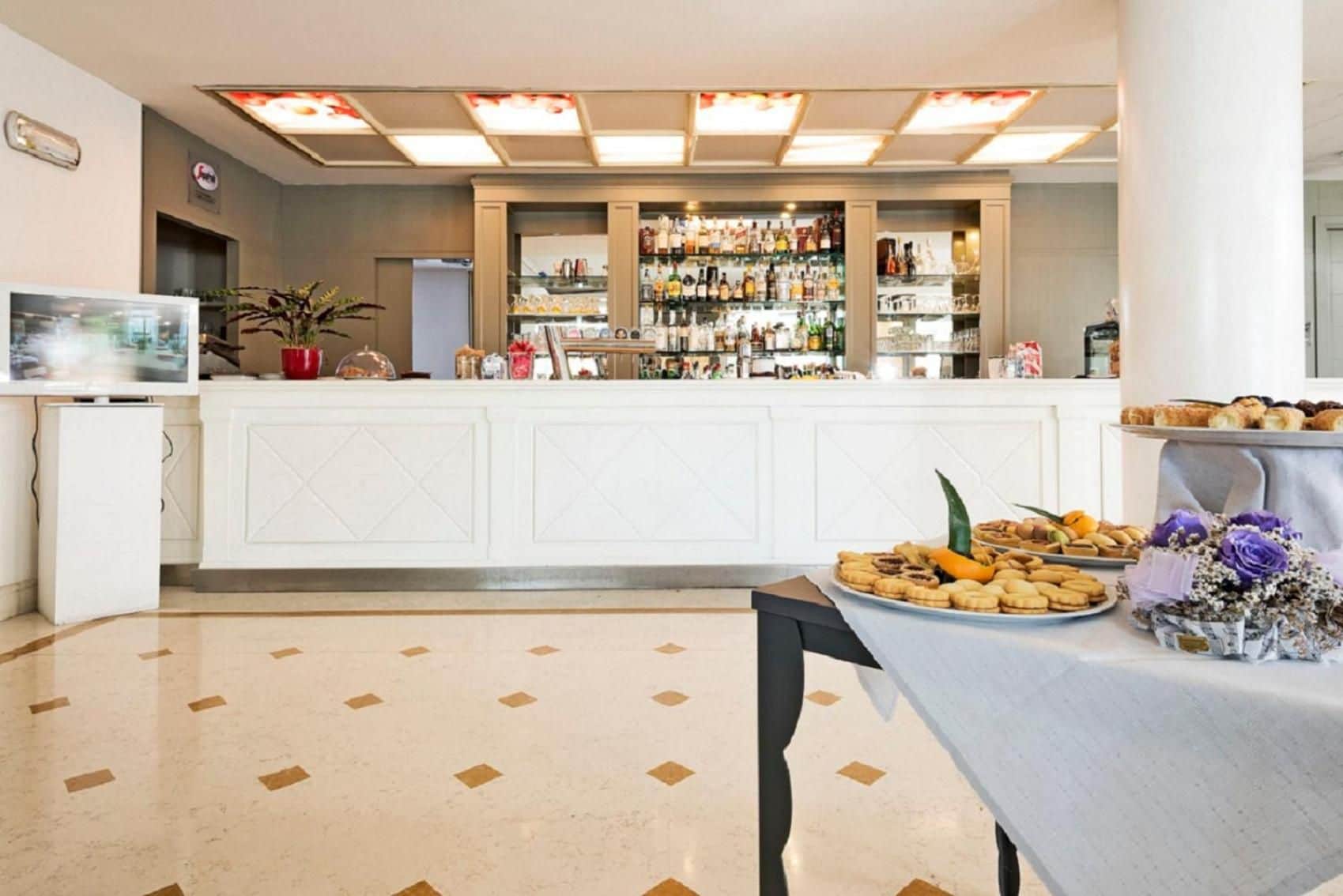 A hotel lobby with a reception desk, a television, a potted plant, and a buffet table with pastries and flowers.