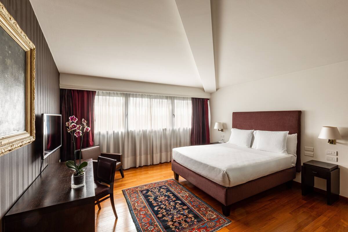 A hotel room with a bed, bedside tables, a desk with a chair, a TV, and a window with curtains.