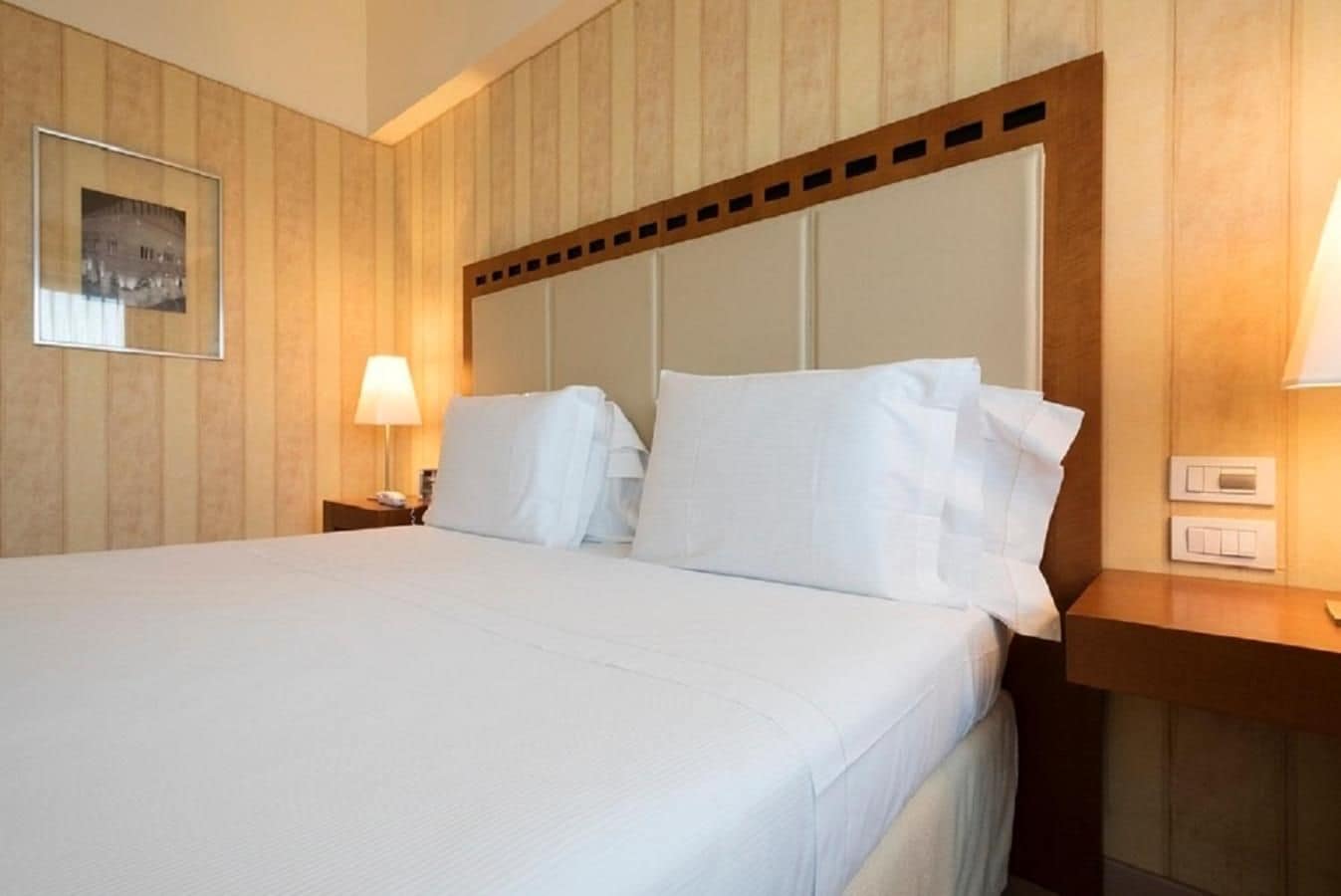 A hotel room with a large bed, wooden headboard, two bedside tables, a nightstand, a lamp, and a framed picture on the wall.