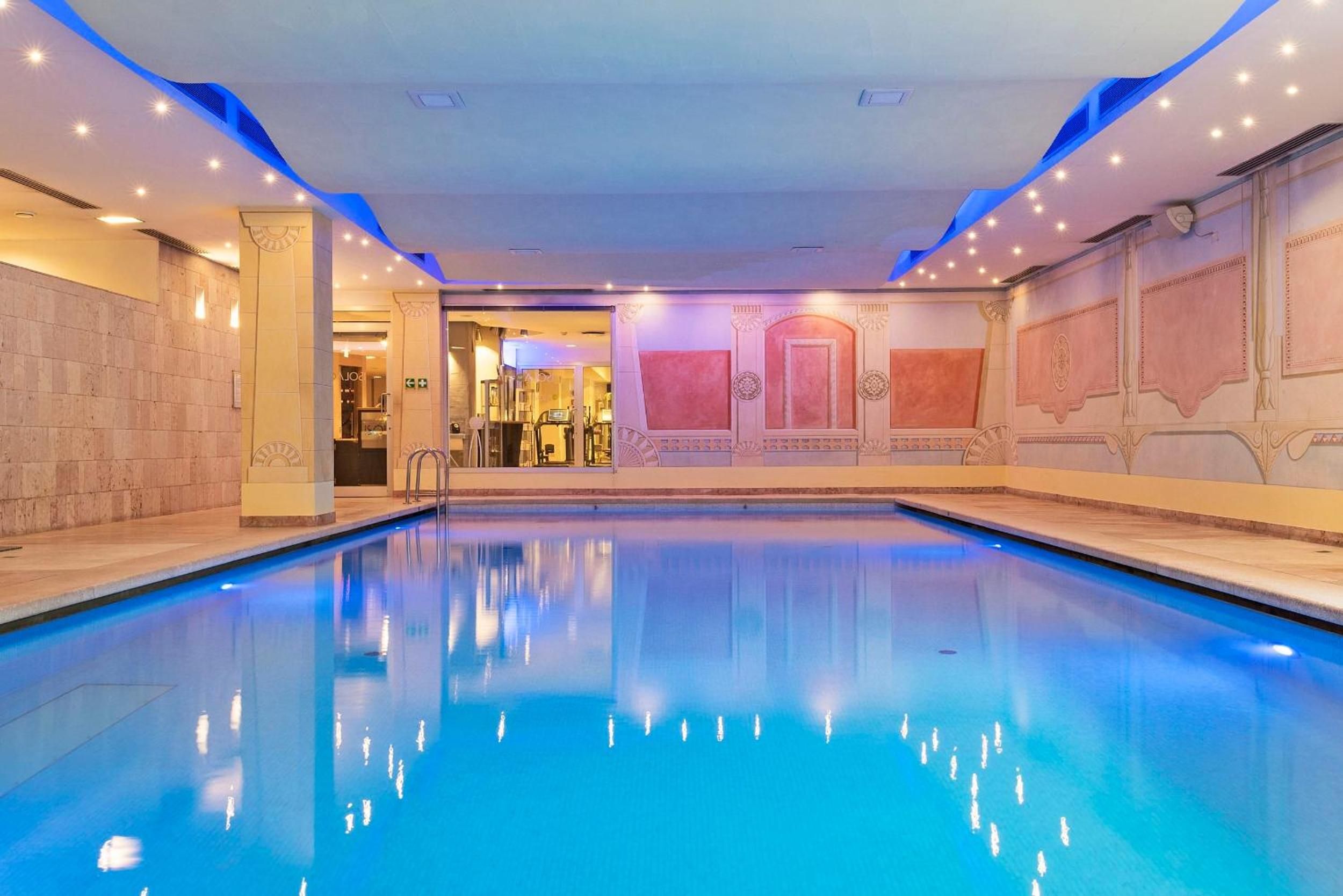 Una gran piscina interior d'aigua cristal·lina i blava, envoltada per parets amb panells decoratius i il·luminació.