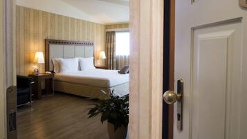 Una habitación de hotel con cama, mesitas de noche, lámparas, una planta en maceta y una puerta.